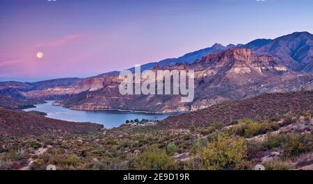 Moon setting down at sunrise over Apache Lake in Superstition Mountains, view from Apache Trail road, Arizona, USA Stock Photo