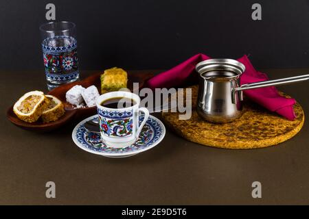 Turkish coffee in cezve with cup, saucer, water glass served with Turkish delight and baclava. Low lighting Stock Photo
