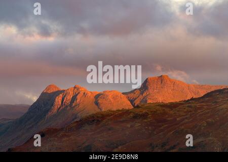 Beautiful red sunlight from the rising sun illuminating the Langdale Pikes mountain range in the English Lake District with dark clouds in sky. Stock Photo