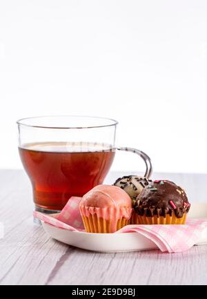 infusion on wooden surface in light tone and white background, accompanied by a plate with mini cup cakes covered in strawberry, dark chocolate and wh Stock Photo