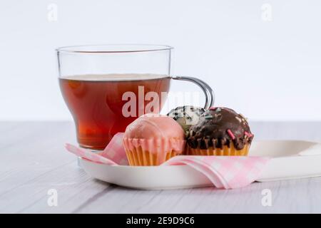 infusion on wooden surface in light tone and white background, accompanied by a plate with mini cup cakes covered in strawberry, dark chocolate and wh Stock Photo