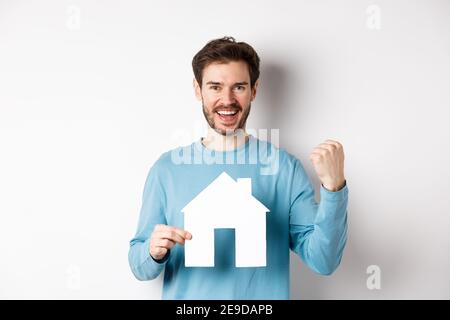 Real estate and insurance concept. Cheerful man buying property and celebrating, saying yes and showing paper house cutout, standing on white Stock Photo