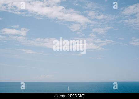 A lone sailboat floats in the distance on calm water against a background of blue sky. The concept of freedom and ease. Blue summer background. Stock Photo