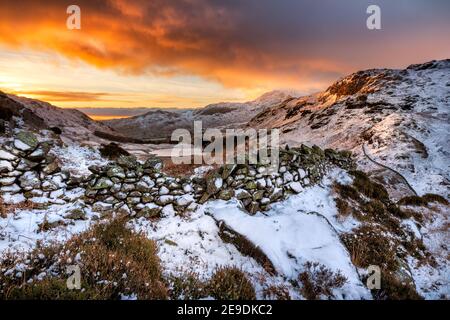 Burning orange clouds in sky at sunrise with snow covered mountains and old stone wall. Lake District, UK. Stock Photo