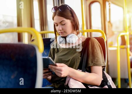 Serious young student girl sitting in a bus and looking at her telephone with serious look. Holding headphones around her neck. Stock Photo