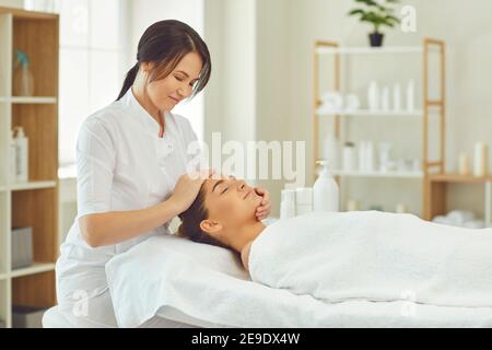 Smiling masseur making procedure of manual relaxing facial massage for young woman Stock Photo
