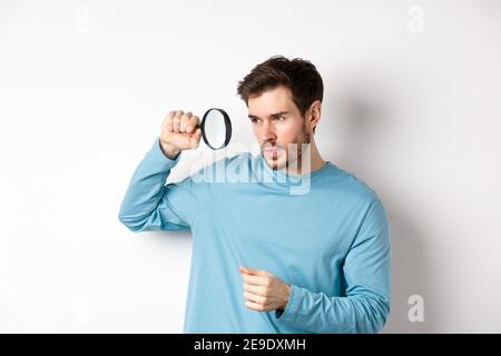 Young handsome man look through magnifying glass with curious face, investigating or searching for something, standing on white background Stock Photo