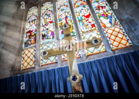 Large brass gold cross with sparkling jewels stood in the sun at front of Church of England chapel with big tall colourful stained glass windows Stock Photo