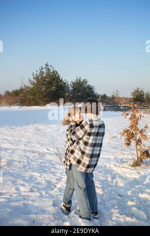 Happy teenage girl and boy, about 16-17 years old, in plaid shirts and jeans, stand in snow hugging and laughing cheerfully and looking at each other. Stock Photo