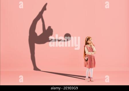 Childhood and dream about big and famous future. Conceptual image with girl and drawned shadow of female rhythmic gymnast on coral pink background. Childhood, dreams, imagination, education concept. Stock Photo