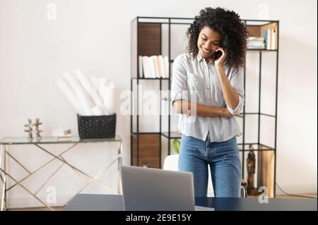 Busy young African American office worker businesswoman, freelancer with Afro hairstyle in casual clothing taking a break from working on a laptop to Stock Photo