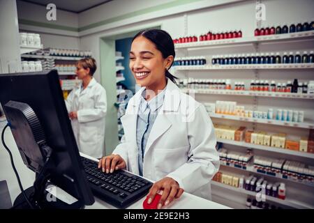 Cheerful young female pharmacist wearing labcoat working on computer in drugstore Stock Photo