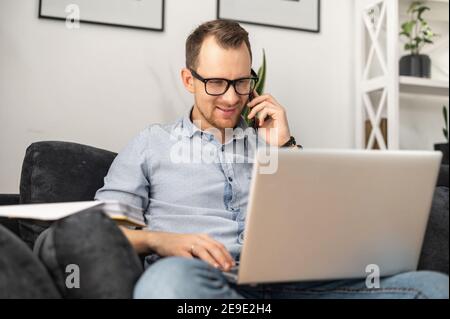 A smiling freelancer guy wearing glasses solving tasks by smartphone working with a laptop from home Stock Photo
