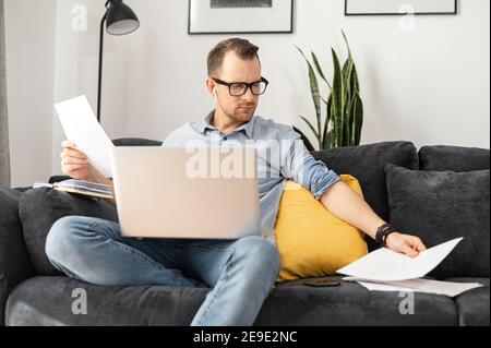 A concentrated businessman wearing eyeglasses working on laptop from home holding documents, doing paperwork, checking balance, preparing report or Stock Photo