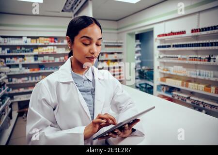 Young female woman working in pharmacy standing behind counter wearing labcoat using digital tablet Stock Photo
