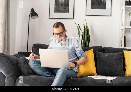 Focused freelancer guy wearing glasses researching business tasks, taking a notes with a laptop at home. A student guy is writing in a notebook Stock Photo