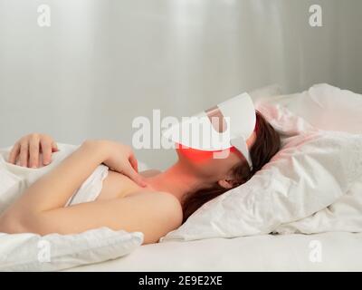 Woman lies in bed with led light therapy facial mask and relax. Home skincare and me time concept. Light rejuvenating mask for facial skin therapy. Photodynamic therapy mask on female face. Copy space Stock Photo