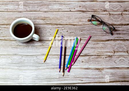 Various colour colouring pencils on a wooden desk or table next to a coffee and glasses Stock Photo