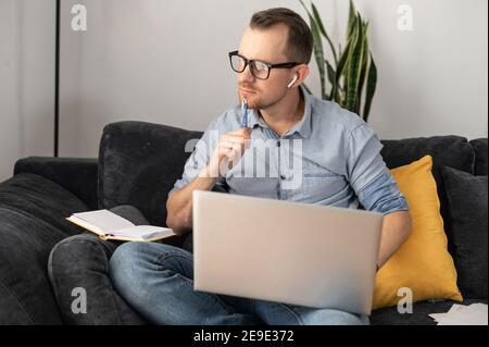 Thoughtful and intelligent entrepreneur looking away sitting on the comfortable couch with laptop. A freelancer wearing glasses works online remotely Stock Photo