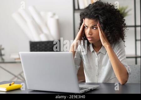 Young African American female office worker feeling stressed, annoyed from the office noise, looking away, touching temples, suffering from severe Stock Photo