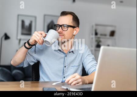 A young man is drinking coffee working with a laptop in the home office. An intelligent guy in glasses with a cup in a hand indoor Stock Photo