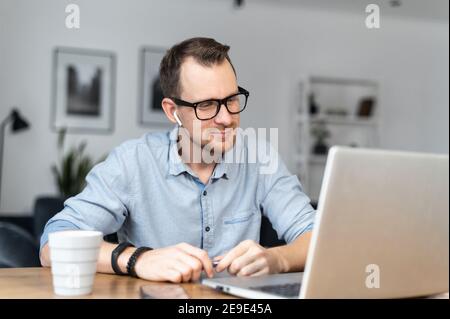 A young man using a laptop in the home office. An intelligent guy in glasses looks at the screen thoughtfully. Remote work concept Stock Photo