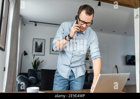 Young successful man is standing near workplace, talking on the phone and check some information on the laptop screen. A freelancer in casual wear is Stock Photo