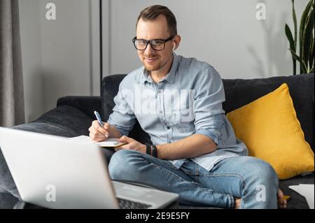 Focused young businessman using laptop for research, writing plan in the notebook, takes notes working online. A guy is work remotely at home sitting Stock Photo