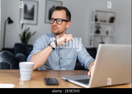 An intelligent young man in glasses and casual shirt sits at the desk with a laptop and looks away lost in thoughts. A freelancer is thinking about Stock Photo