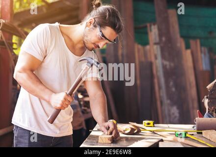 A young male carpenter builder working overall equals a wooden bar with a milling machine in the workshop, in the background wooden Stock Photo
