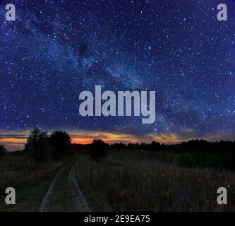 Starry summer night landscape - Milky Way over countryroad on meadow. Night view of natural glowing stars on dark blue sky. Narrow strip of bright sun Stock Photo