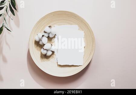 Summer still life scene with an olive branch and shadows on a delicate pink background. A blank sheet of paper with cotton flowers lies on a bamboo pl Stock Photo