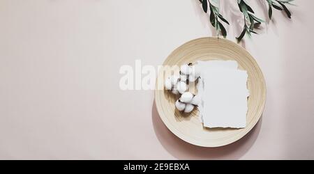 Summer still life scene with an olive branch and shadows on a delicate pink background. A blank sheet of paper with cotton flowers lies on a bamboo pl Stock Photo