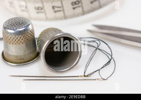 Two sewing thimbles, scissors,  centimeter and two needles with black thread lie on a white background