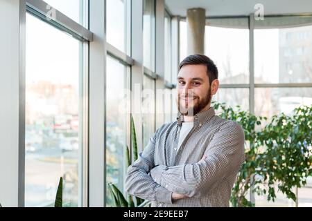 Smiling caucasian man standing near full length window in office alone. Successful bearded young smart man crossed arms. Handsome businessman in Stock Photo