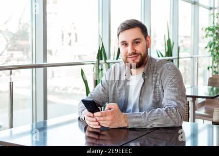 Smiling caucasian man texting message on smartphone in cafe alone near window Stock Photo