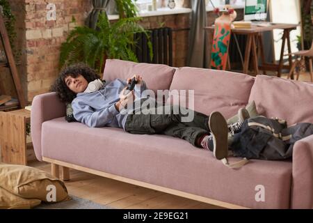 Guy lying on the sofa with joystick and playing video game during his leisure time in the room Stock Photo