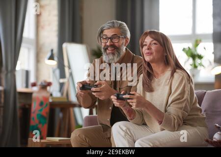 Mature couple sitting on sofa with joysticks and playing video games together at home Stock Photo