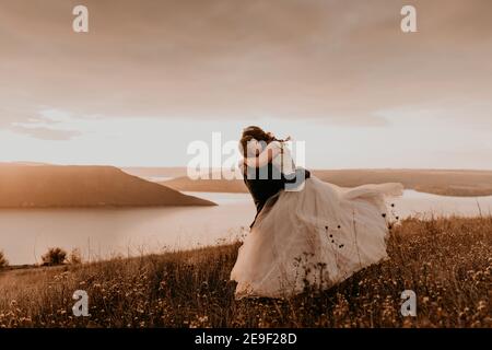 loving couple wedding newlyweds in white dress and suit hug kissing whirl on tall grass in summer field on mountain above the river. sunset. Stock Photo