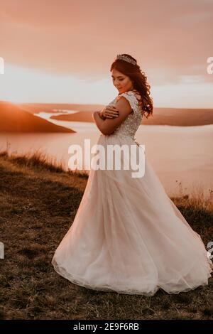 young brunette bride in white wedding dress with a crown on her head stands on cliff against the background of the river and islands After sunset Stock Photo