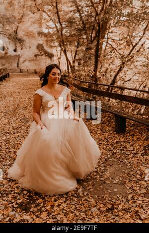 woman bride in white wedding dress with hairstyle makeup and crown on her head walks through autumn forest on fallen orange leaves Stock Photo