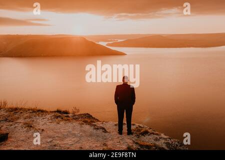 man in formal suit groom stands on mountain holds his hands in pockets. fiery orange sunset sunset rays through clouds. panoramic view from cliff to Stock Photo