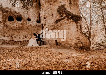 wedding couple in love man and woman sitting on a bench under rock monastery bakota in autumn forest against background of stone rocks. groom suit and Stock Photo