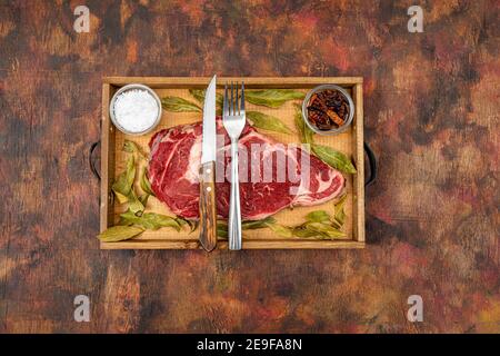 Raw beef ribeye on a round slate plate with glass containers containing peppercorns, coarse salt and cayenne chillies on an old wooden table in brown Stock Photo