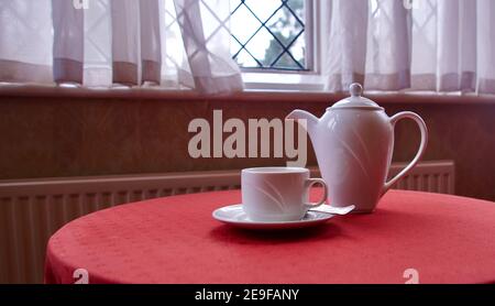 White china coffee pot and cup and saucer on red table below window