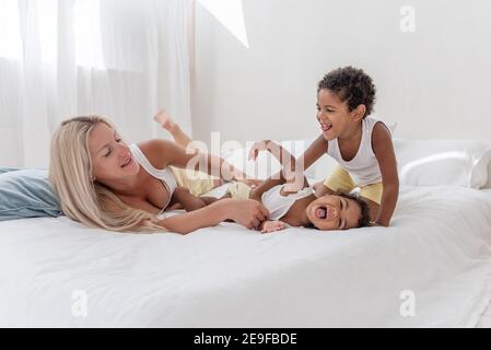 Young Caucasian blond mother plays, lies on white bed with African American daughter son. Happy multi ethnic family morning. Woman hugs, strokes child Stock Photo