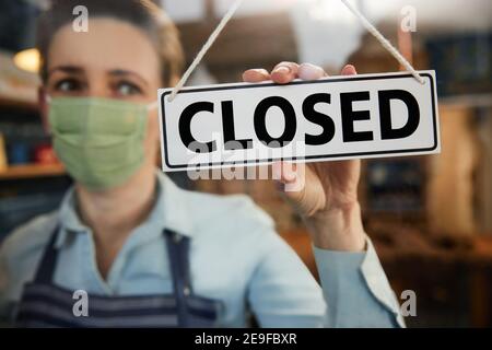 Female Owner Of Small Business Wearing Face Mask Turning Round Closed Sign During Health Pandemic Stock Photo