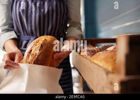 Sales Assistant In Bakery Putting Freshly Baked Organic Sourdough Bread Loaf Into Sustainable Paper Bag Stock Photo