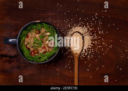 top view of vegan quinoa bowl on wooden table with rocket salad, olives, peas, tomatoes. rustic healthy homemade dish with wood spoon Stock Photo