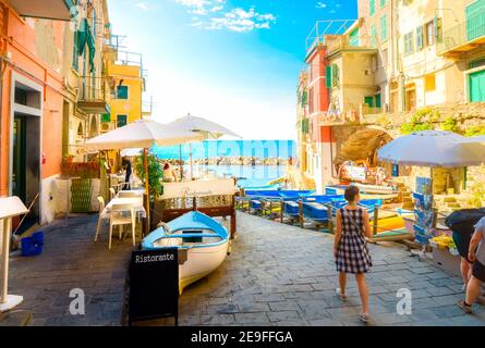 A young woman in a plaid dress walks towards the boat launch past sidewalk cafes in the colorful Cinque Terre village of Riomaggiore, Italy. Stock Photo